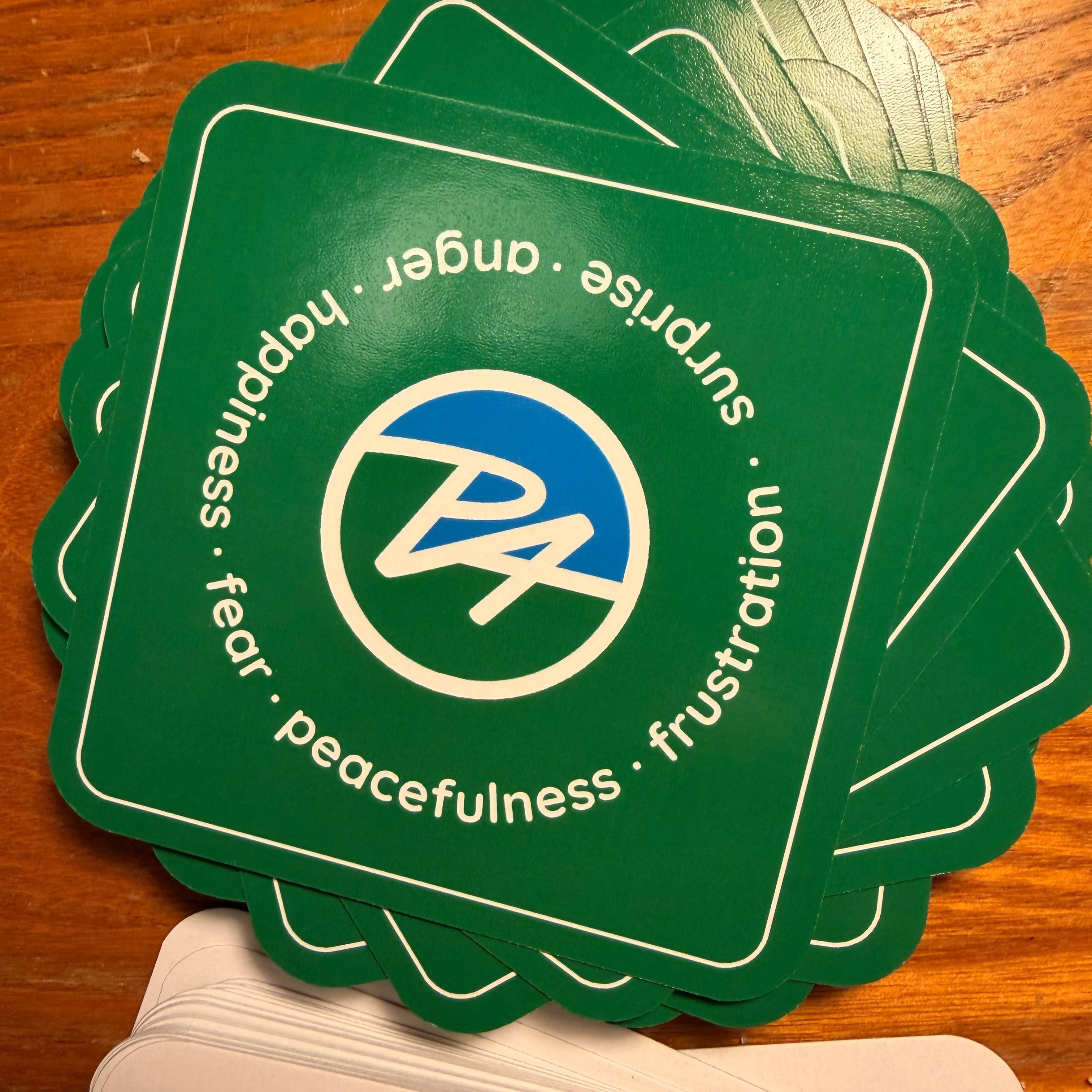 A stack of green square cards with rounded corners on a wooden surface. The top card shows a white border, a blue and white circular logo in the center, and white text around the border reading 'anger, happiness, fear, peacefulness, surprise, frustration'.