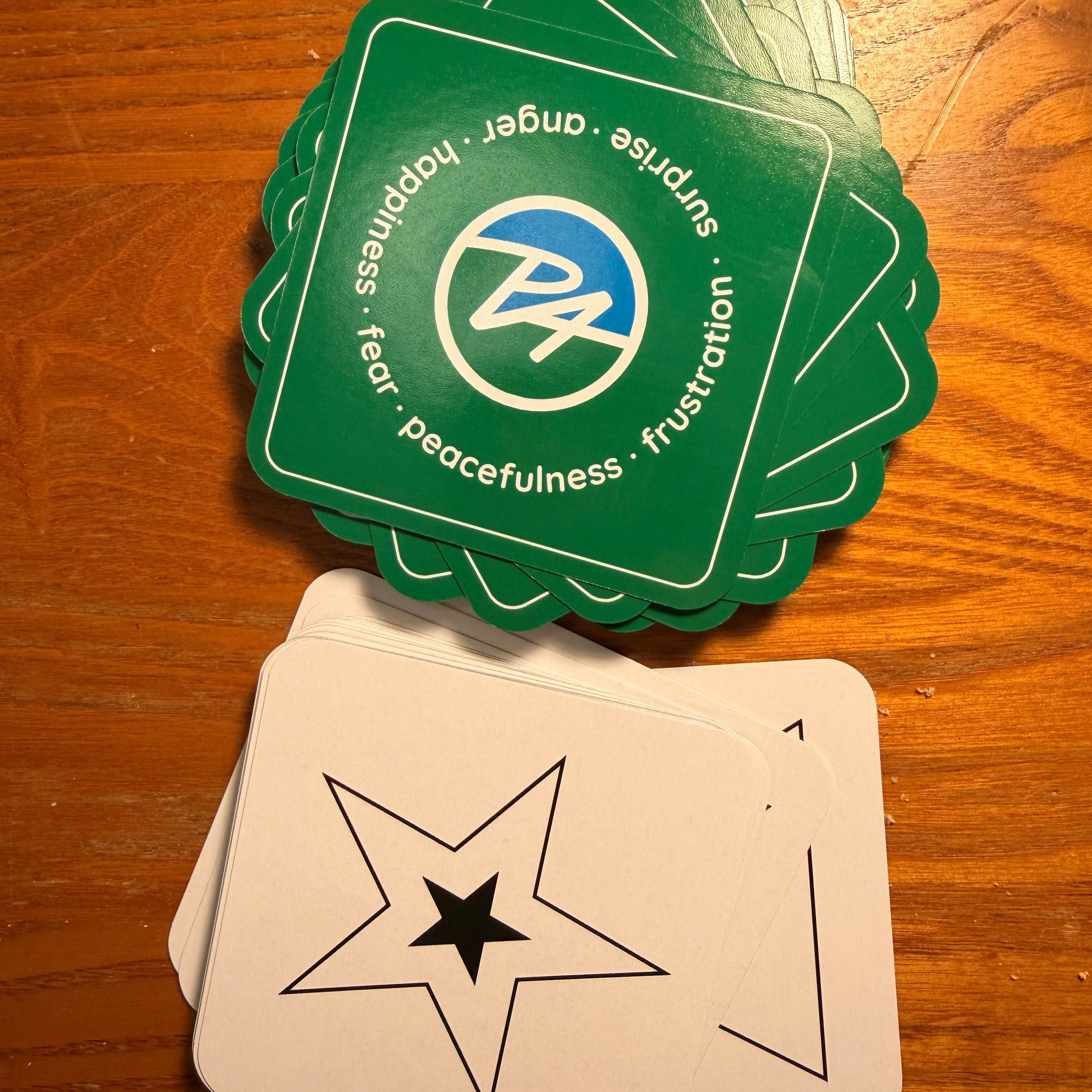 A deck of cards laid on a wooden table, with one face-up card showing a black star outline on an off-white background and a second stack of green scalloped cards showing a blue and white circular logo with text around the edge.