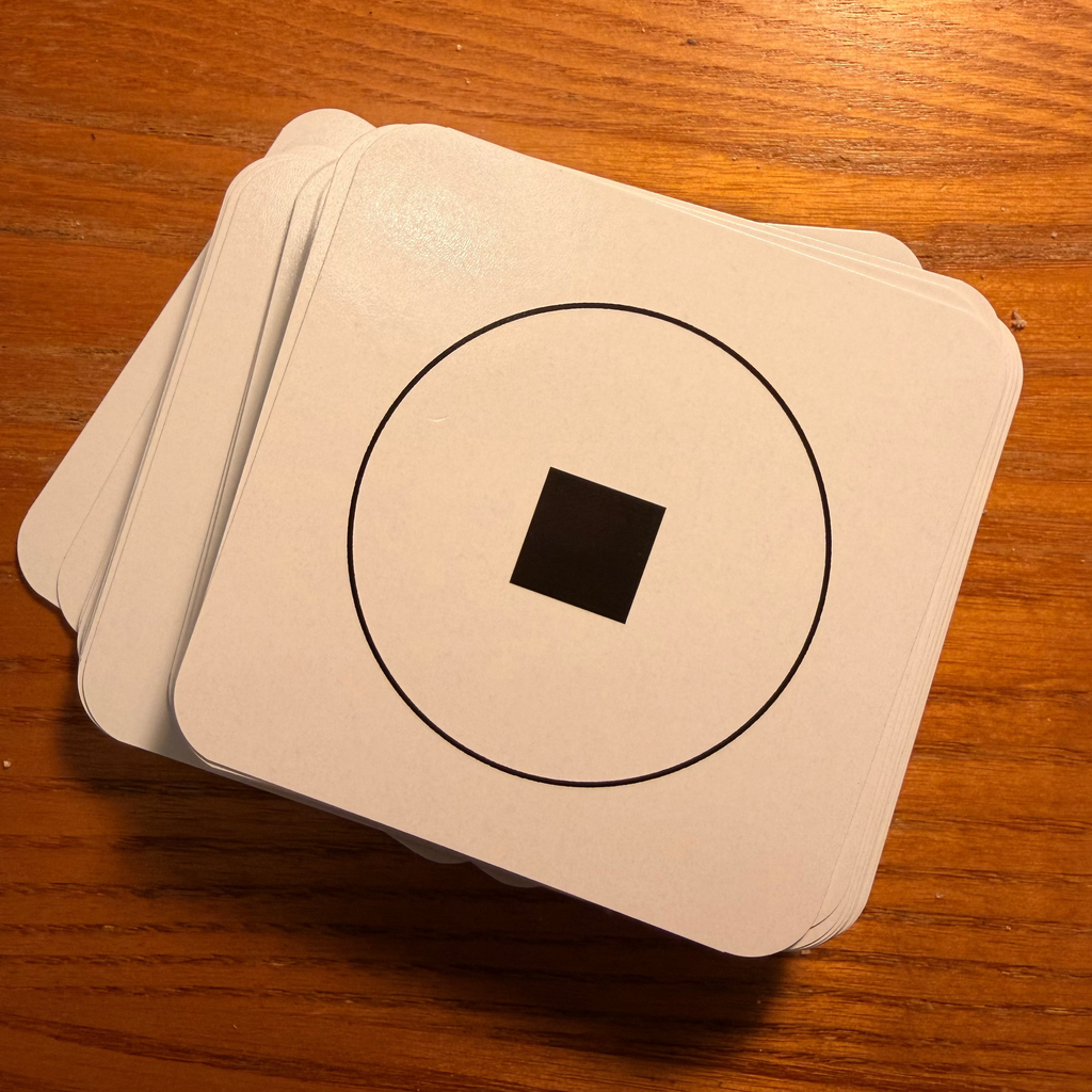 A stack of square white cards with a minimalist black geometric design (a square inside a circle) resting on a warm brown wooden table.